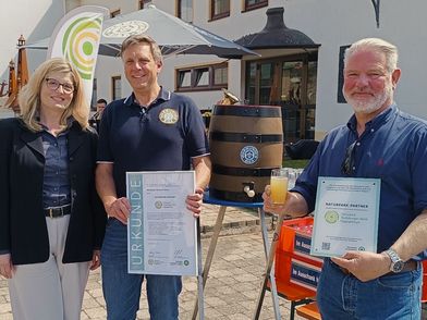 Die Urkunde für die Naturparkpartnerschaft wird überreicht. Es sind drei Personen auf dem Bild, eine Frau und zwei Männer.