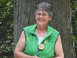 Das Bild zeigt Naturparkführerin Marie-Luise Eickmeier-Ehrlich in grüner Weste und bloßen Armen. Sie lacht fröhlich, hat kurzes ergrauendes Haar und steht vor einem dicken Baum.