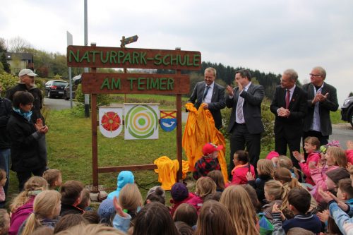 Kinder sitzen im Halbkreis um neues Schild. Erwachsene stehen daneben und klatschen.