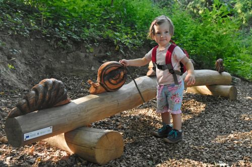 Kleines Mädchen Steht vor einem geschnitzten Baumstamm mit geschnitzter Schnecke.